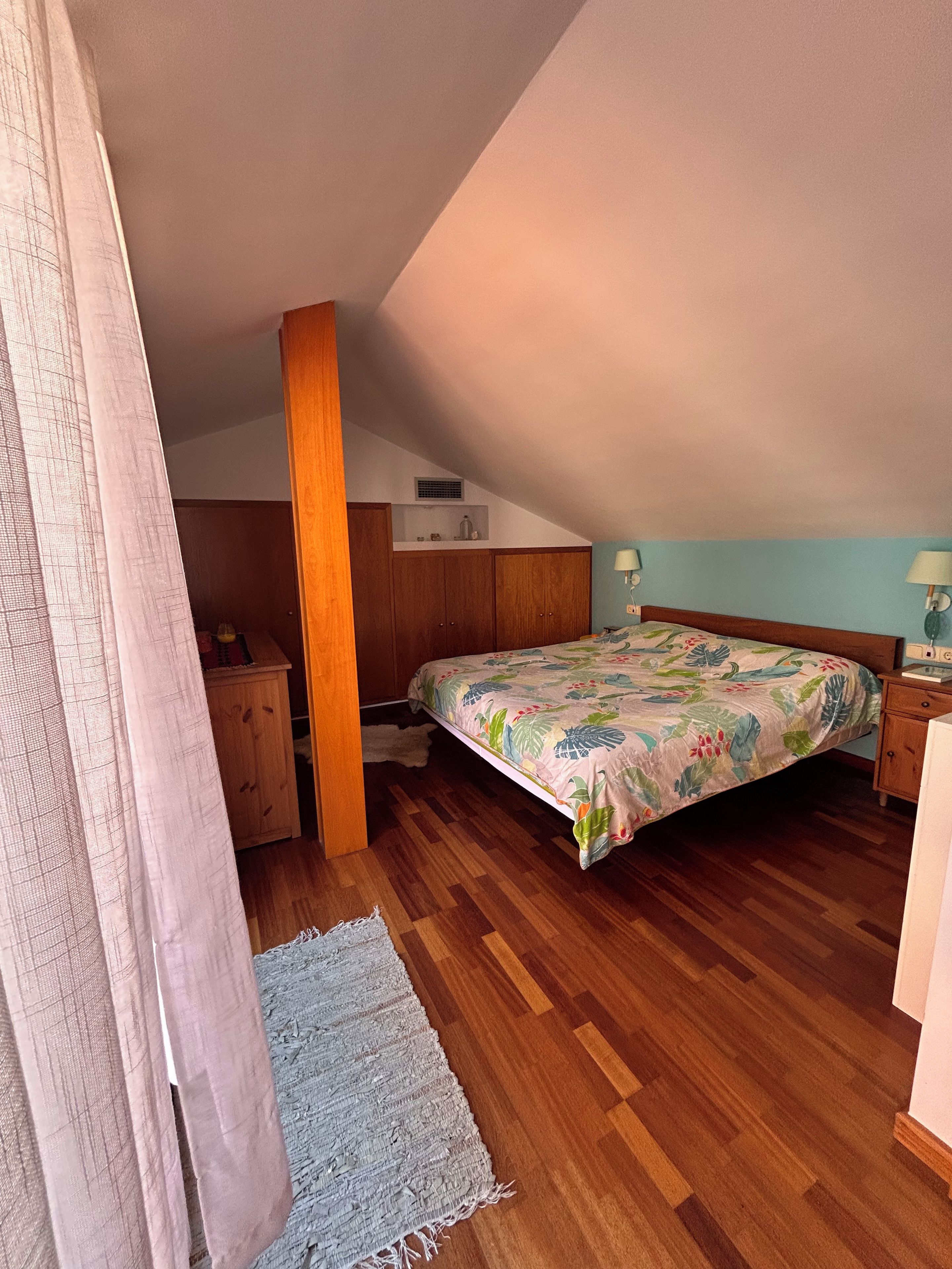 A bedroom with a wooden floor, a bed with a floral bedspread, a nightstand, a wooden wardrobe, and a small shelf with items.