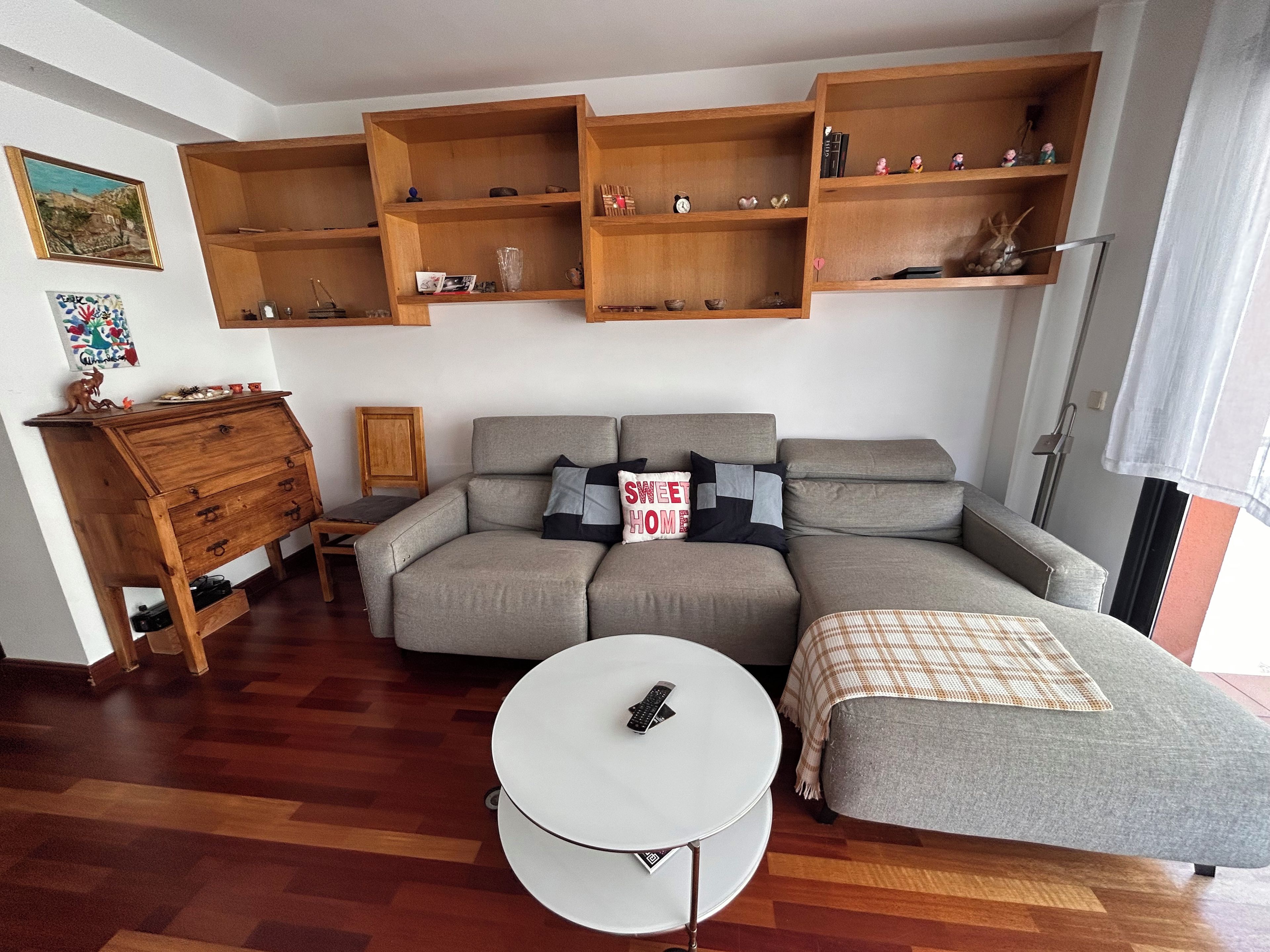 A living room with a sectional sofa, a small round table, wooden shelves above, and a wooden cabinet.