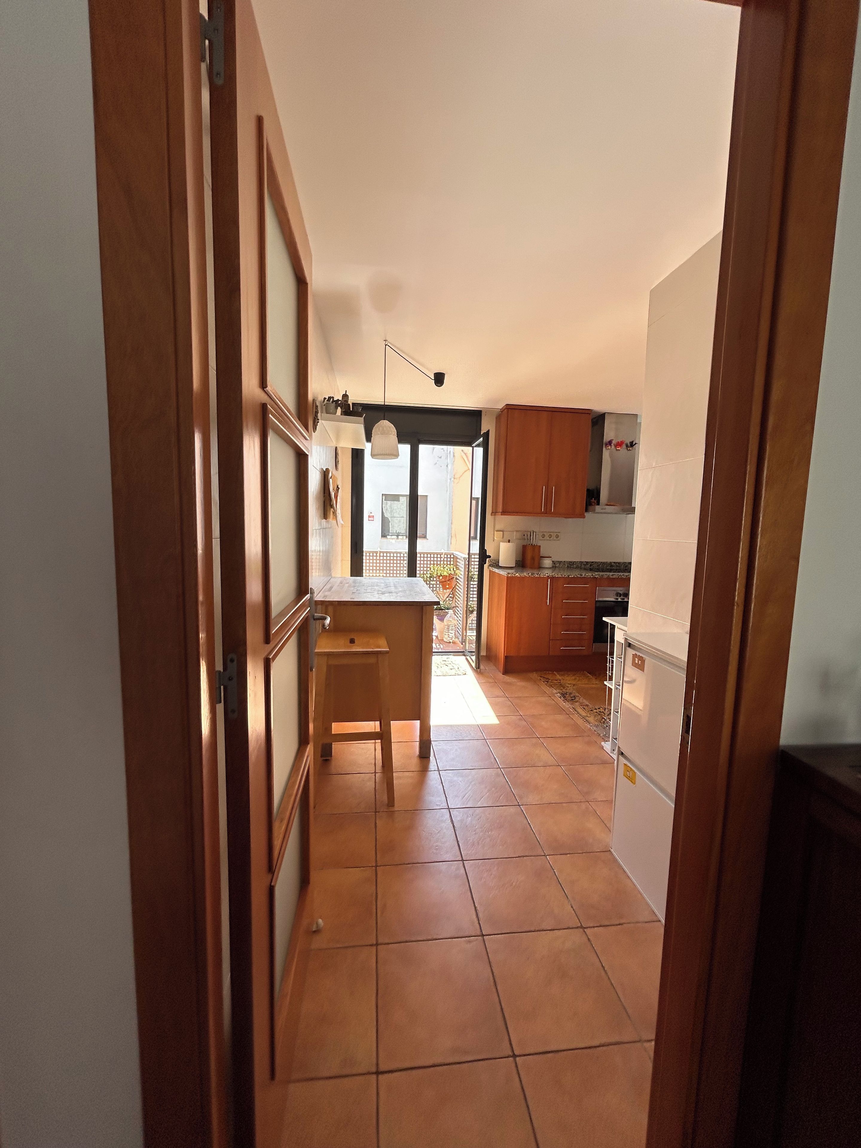A narrow hallway leading to a kitchen with wooden cabinets, a small stool, and a tiled floor.