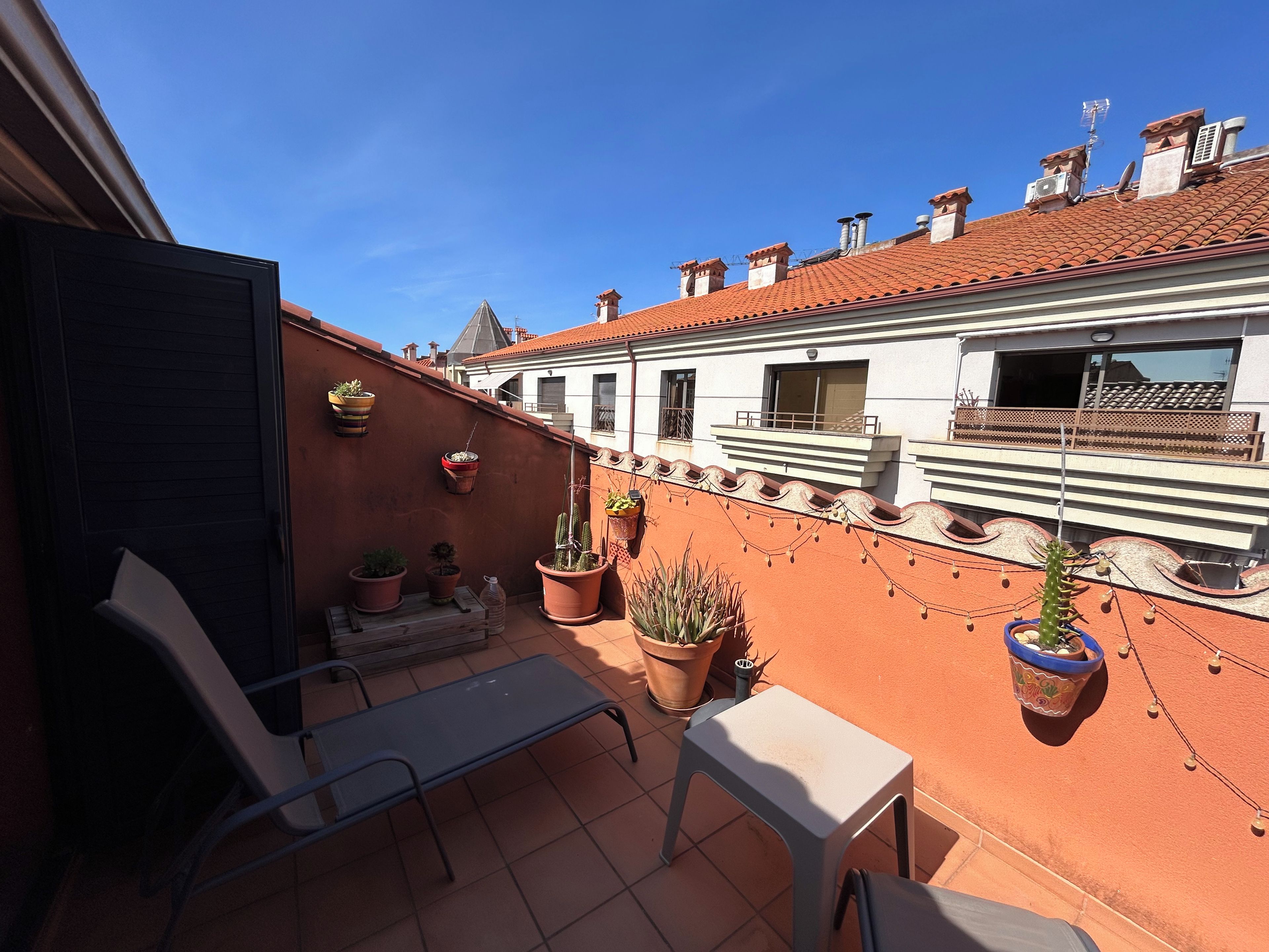 A rooftop terrace with potted plants, a metal table, and a bench.