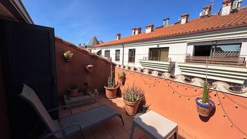A rooftop terrace with potted plants, a metal table, and a bench.