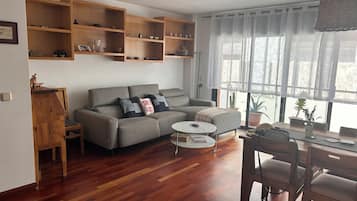 A modern living room with a grey sofa, wooden shelves, a dining table with chairs, and a large window with sheer curtains.