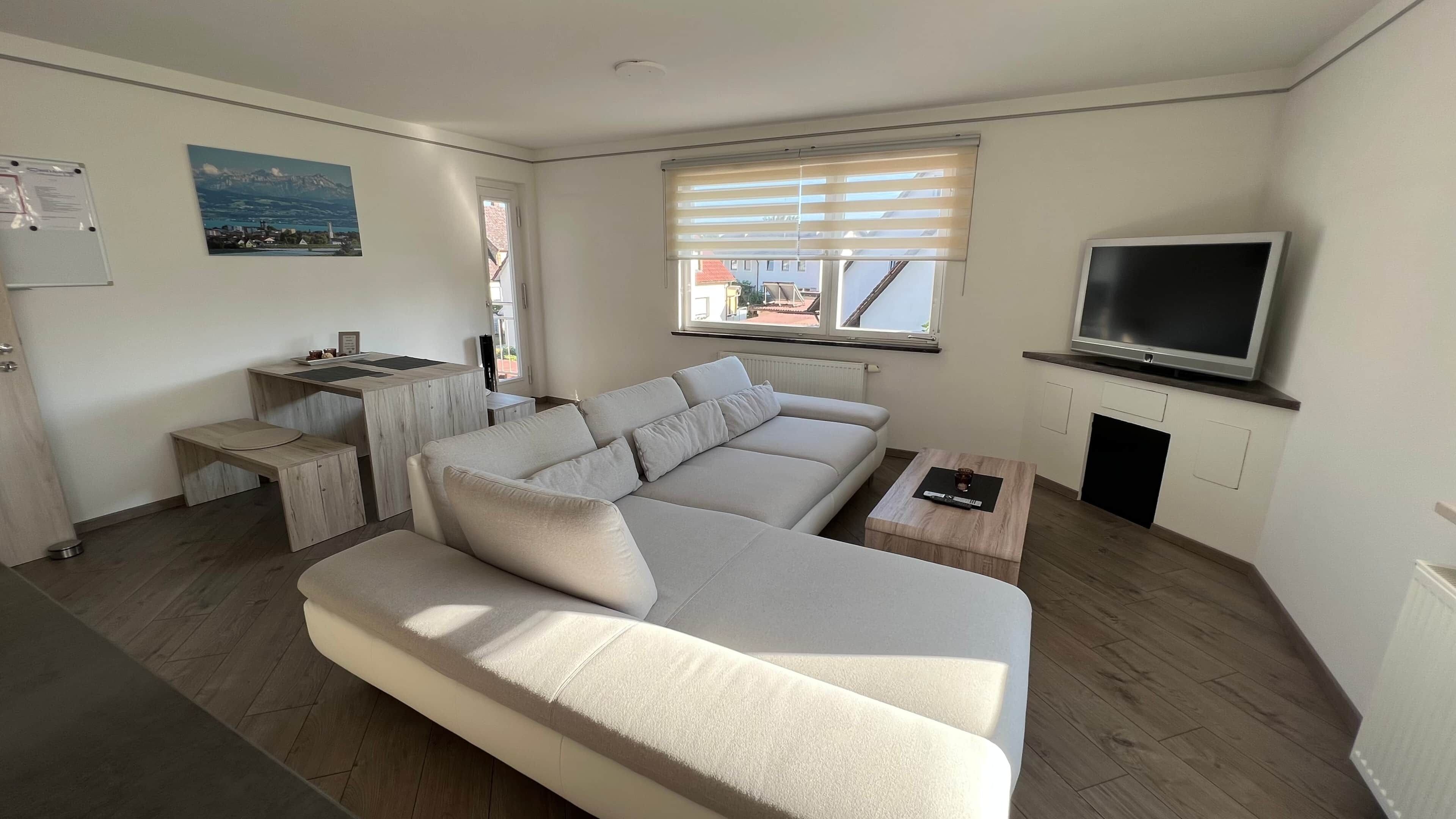 Ein modernes Wohnzimmer mit einer Couch, einem kleinen Tisch, einem Fernseher und einem Fenster mit Jalousien.