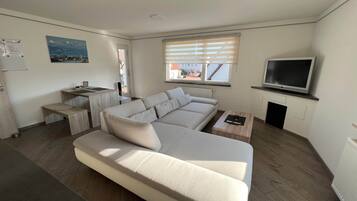 Ein modernes Wohnzimmer mit einer Couch, einem kleinen Tisch, einem Fernseher und einem Fenster mit Jalousien.
