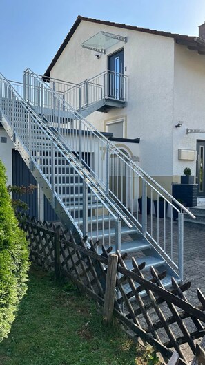 A building with a metal staircase leading to a door, a glass awning, and a wooden fence. - David´s Apartment (Meckenbeuren)