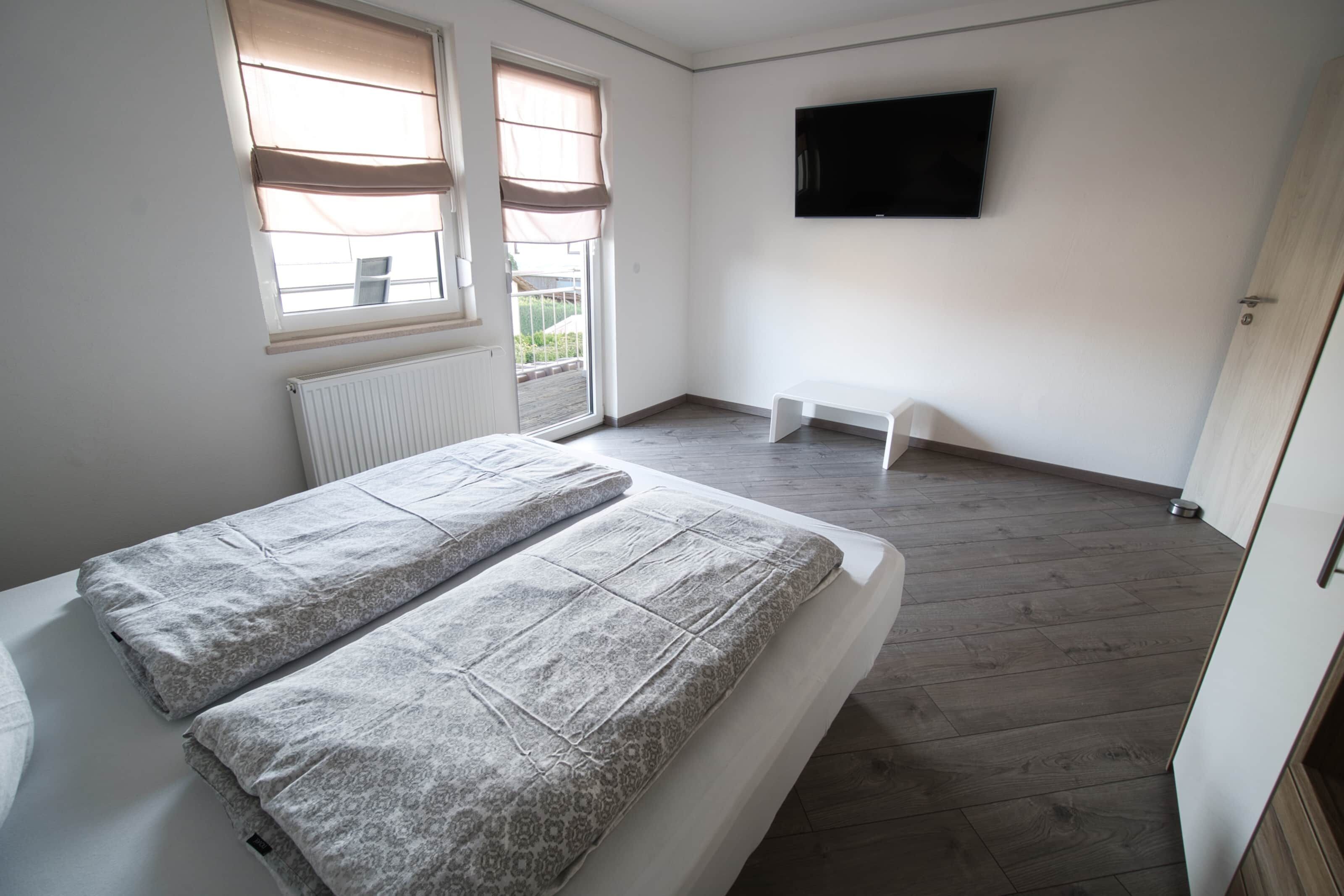 A bedroom with a bed, a radiator, a window with blinds, a television on the wall, and a door.