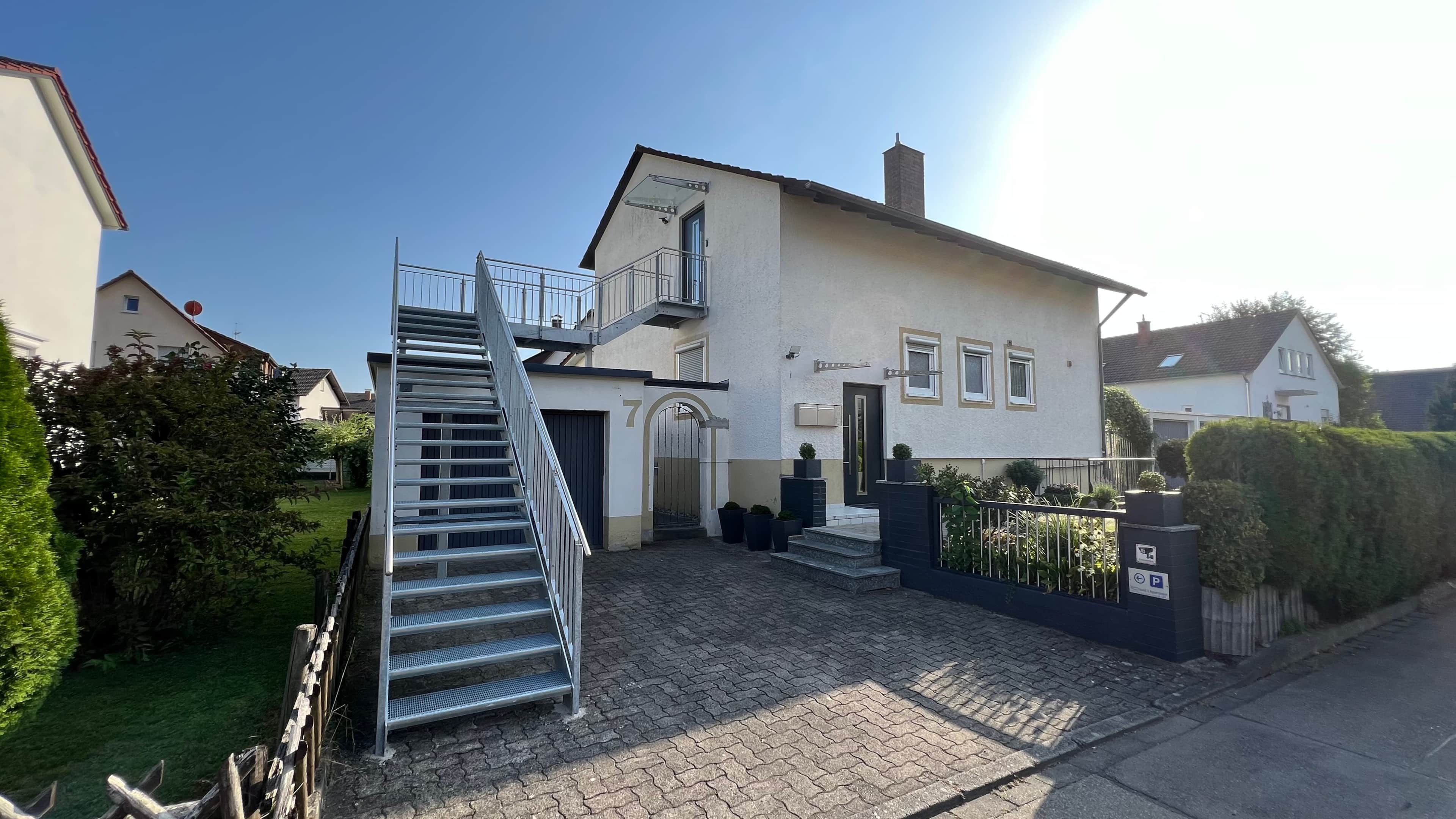 Ein zweistöckiges Haus mit einer Treppe zum Eingang, einer Einfahrt mit Tor und einem gepflegten Rasen.