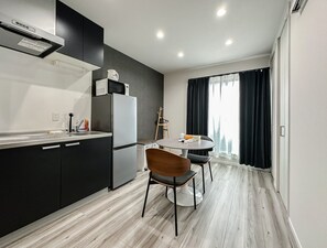 A modern kitchen with a refrigerator, microwave, and a kettle on the counter. There is a dining table with chairs and a window with curtains. - HANASTAY IWA (Osaka)