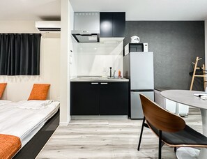 A compact hotel room with a bed, a small kitchenette, a dining area, and a wooden chair. - HANASTAY IWA (Osaka)
