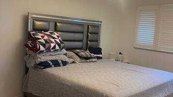 A bedroom with a bed, pillows, a nightstand, a lamp, and a window with blinds.