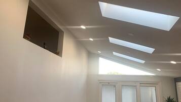 A room with a skylight, recessed ceiling lights, a window with blinds, and a small shelf with decorative items.