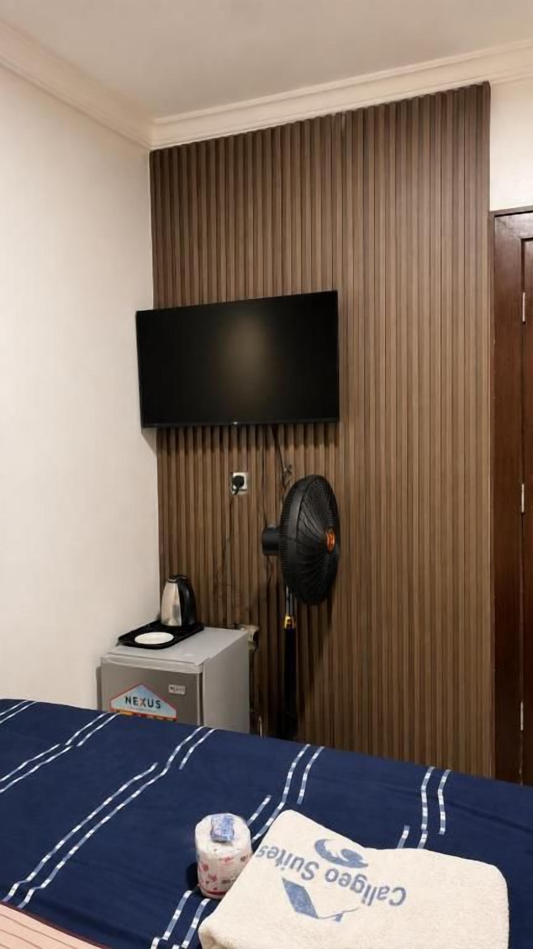 A hotel room with a bed, a flat-screen TV mounted on the wall, a standing fan, and a small table with a kettle.