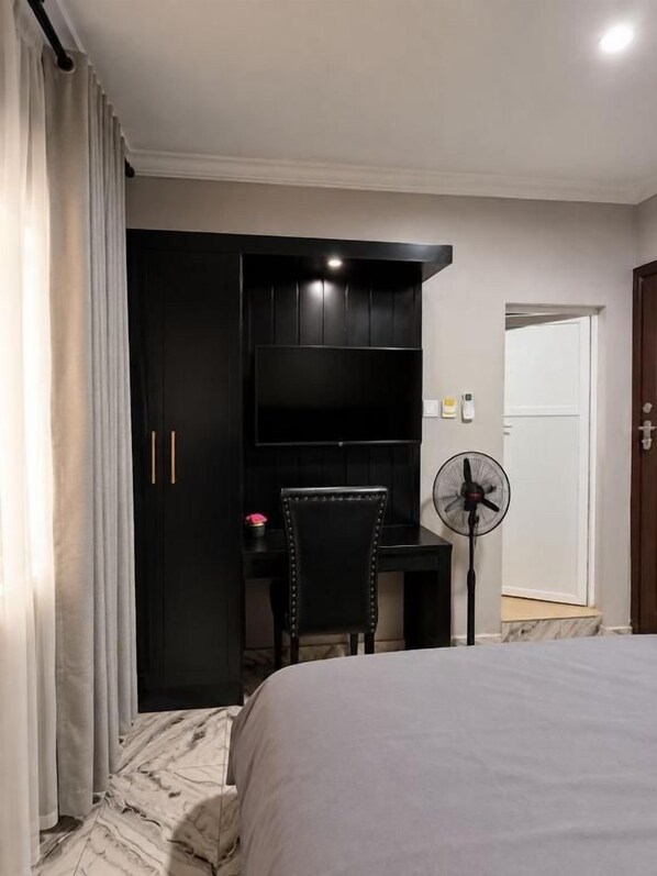 A bedroom with a bed, a desk, a chair, a fan, and a wardrobe. - Caligeo Suites II (Ibadan)