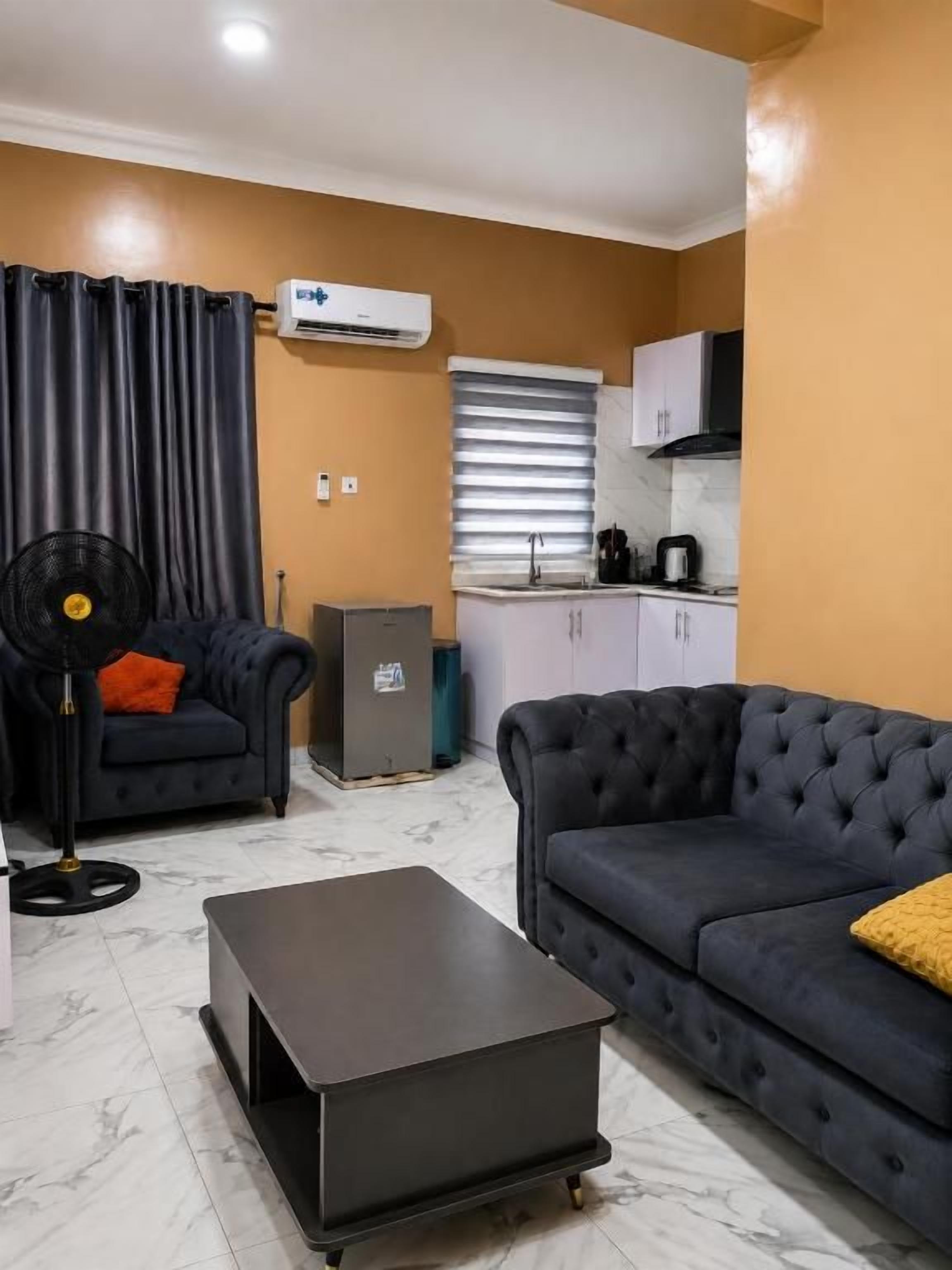A living room with a sofa, a coffee table, and a fan. There is a kitchen area with cabinets and a sink in the background.