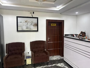 A reception area with two brown upholstered chairs, a wooden door with a sign, and a counter with a person behind it. - Caligeo Suites II (Ibadan)