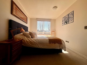 A bedroom with a bed, bedside table, lamp, and a window with a view of trees. - COZY, WARM, FLEXIBLE, CENTRAL, 2 bedrooms, 2 bathrooms, 3 living areas + parking (Puerto Varas)