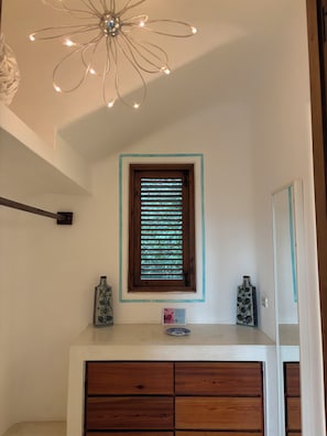 Casa de banho com toucador de madeira, um vaso decorativo, iluminação embutida na parede e janela com persianas.