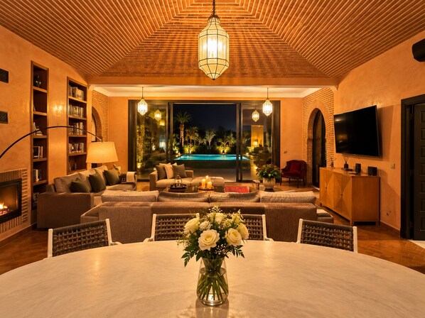 A modern living room with a fireplace, a pool view, and a dining area with a vase of flowers.