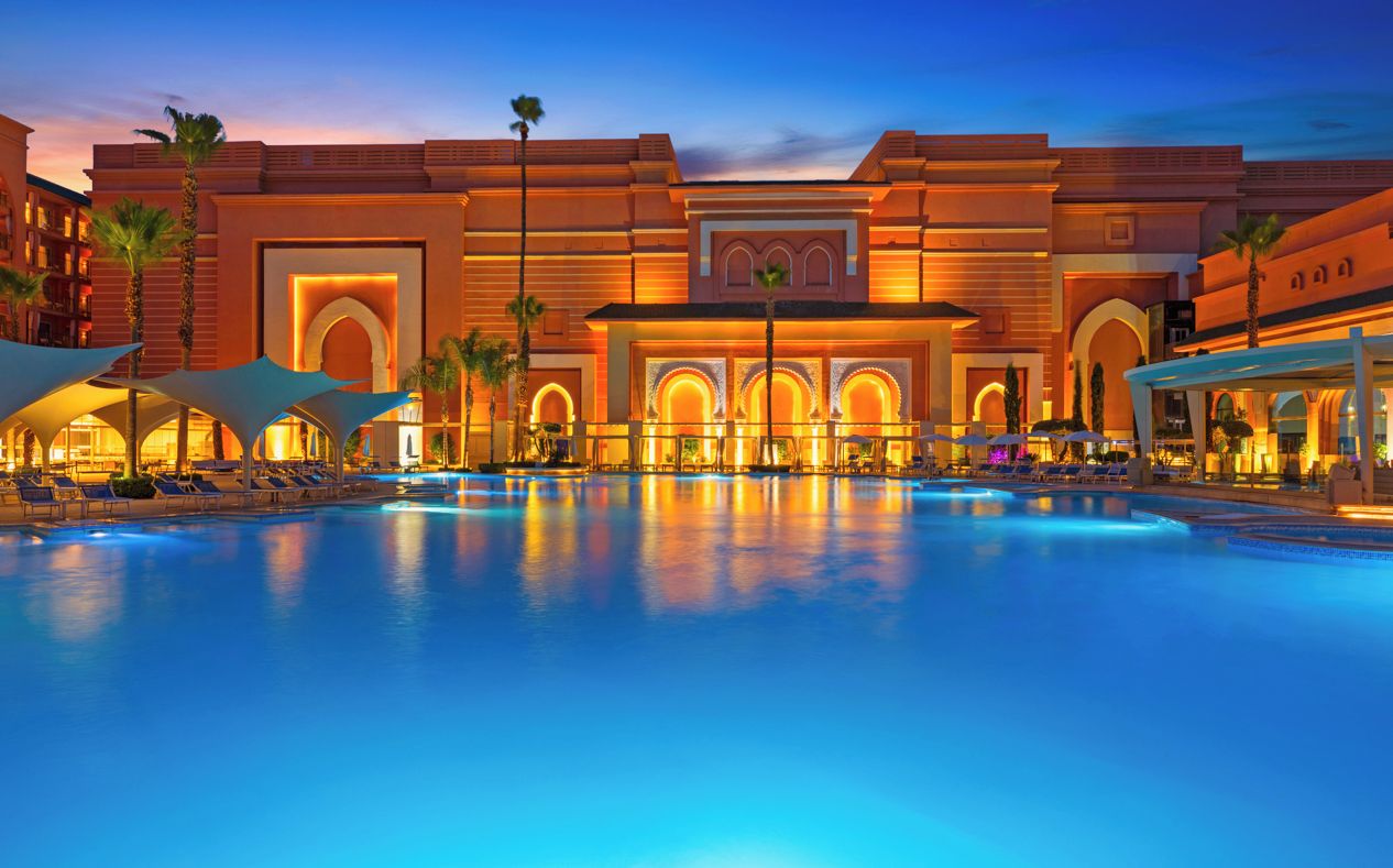 Un hotel con una amplia piscina, palmeras y una imponente entrada con arcos.