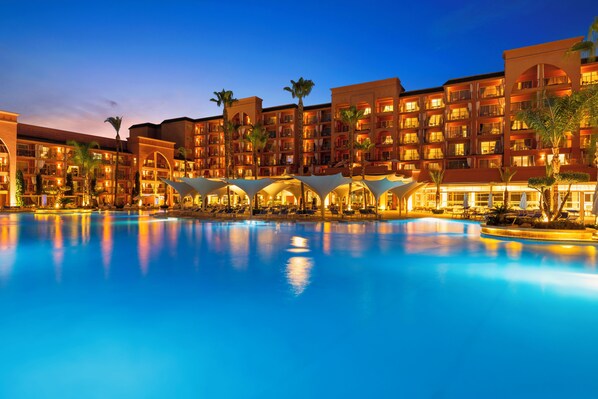 A hotel with a large swimming pool, multiple balconies, and palm trees.