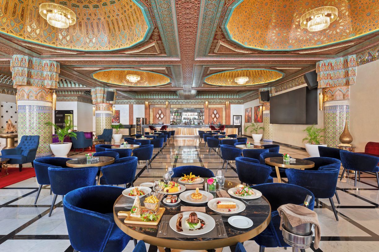 Un hall d’hôtel spacieux, avec une longue table dressée pour le dîner, entourée de chaises bleues et agrémenté de décorations ornementales au plafond.