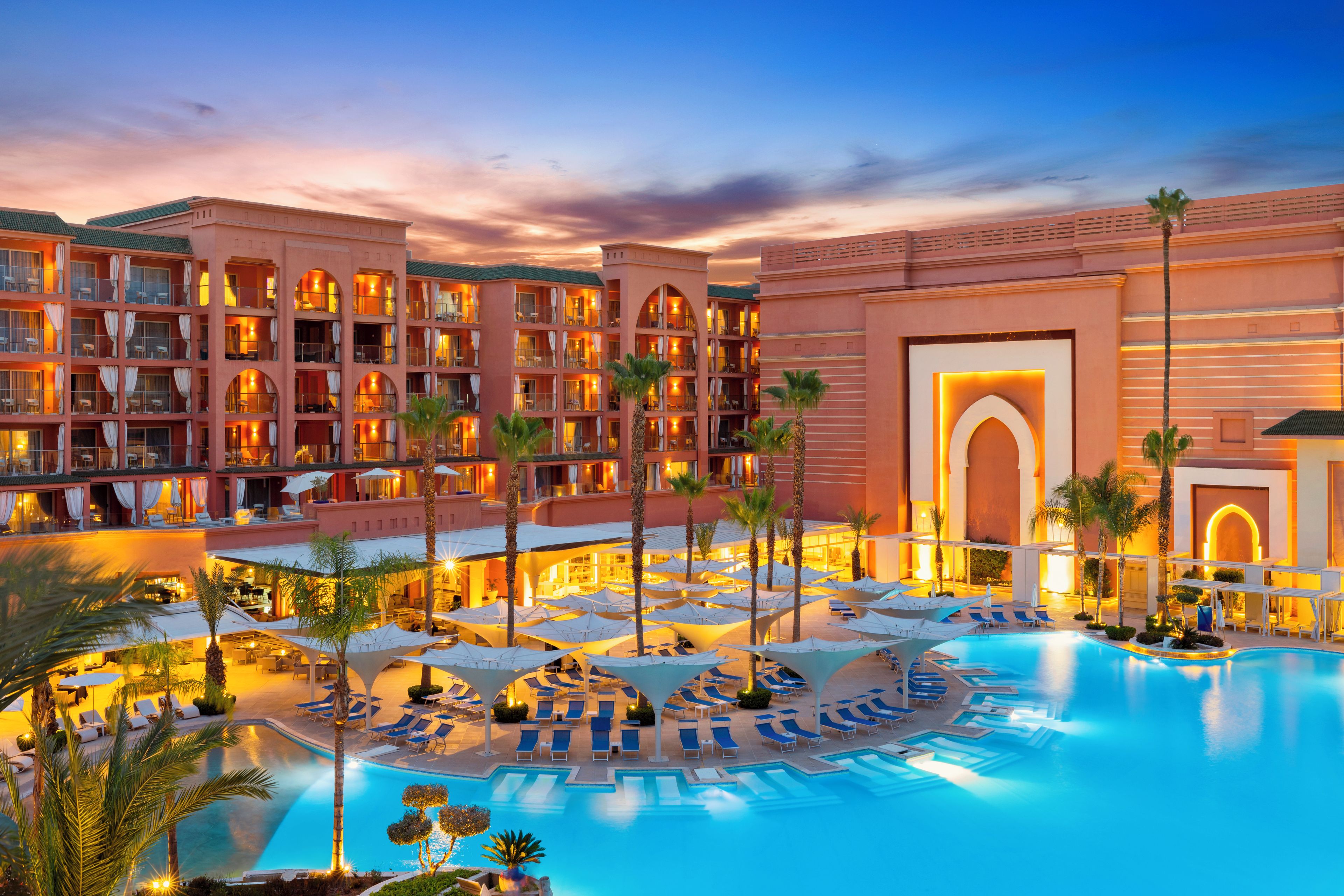 Un hotel de varios pisos, con zona de piscina y tumbonas, y palmeras.