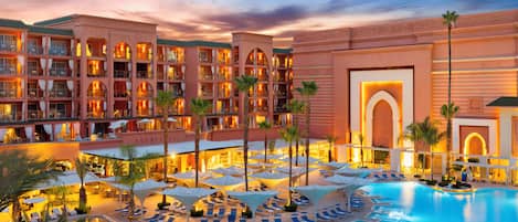 A hotel with multiple floors, a pool area with sun loungers, and palm trees.