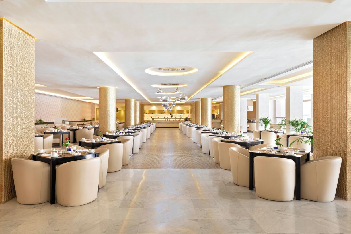 Un vaste espace de restauration d’hôtel, doté de nombreuses tables et chaises, d’un passage central et d’un design de plafond moderne.
