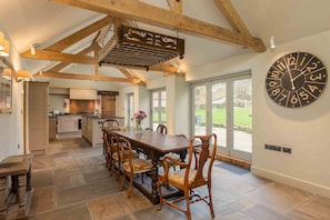 A spacious dining area with a large wooden table and chairs, a kitchen in the background, and a large wall clock. - Scugdale -  a holiday cottage that sleeps 4 guests  in 2 bedrooms (Hartoft)