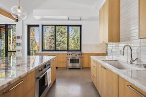 Eine moderne Küche mit Holzausbau, einem Edelstahlbackofen, einem großen Fenster mit Blick auf Bäume und einer Marmorarbeitsplatte.