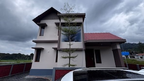 Un edificio de dos pisos con techo de tejas rojas, un pequeño árbol en la entrada y un coche estacionado frente.