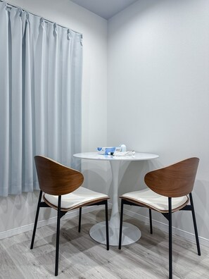 A small room with a round table and two chairs, a curtain, and a small table with a bottle and a box. - HANASTAY HIKARI (Osaka)