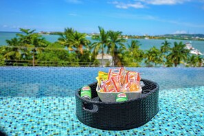 Un cadre agréable au bord de la piscine, avec un panier en osier contenant des collations et des boissons, une vue dégagée sur la mer et des palmiers en arrière-plan.