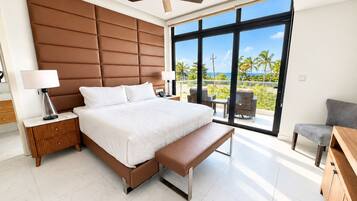 A modern bedroom with a large bed, a ceiling fan, a balcony with outdoor seating, and a view of the ocean.