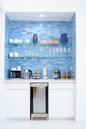 Une cuisine avec un mur recouvert de carreaux bleus, une machine à café, une bouilloire et divers objets en verre disposés sur des étagères.