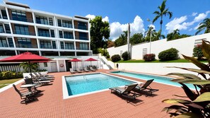 Ett hotellområde med en pool, solstolar och parasoller, en klarblå himmel och en modern byggnad i bakgrunden.