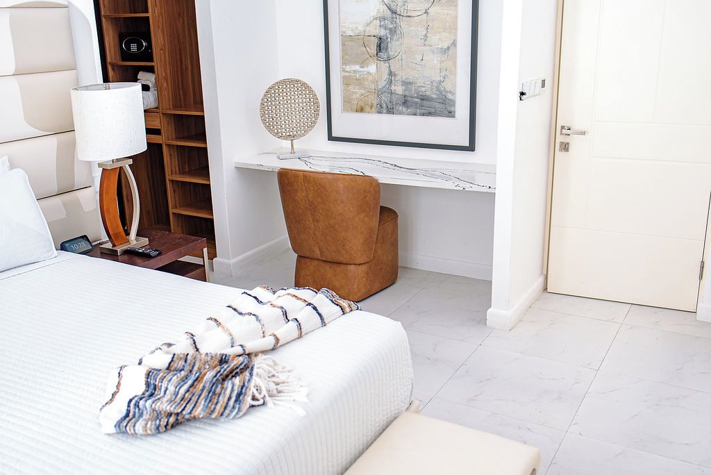 A hotel room with a bed, a desk, a chair, a lamp, a shelf, and a door.