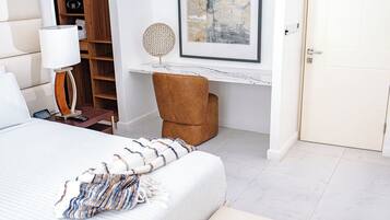 A hotel room with a bed, a desk, a chair, a lamp, a shelf, and a door.