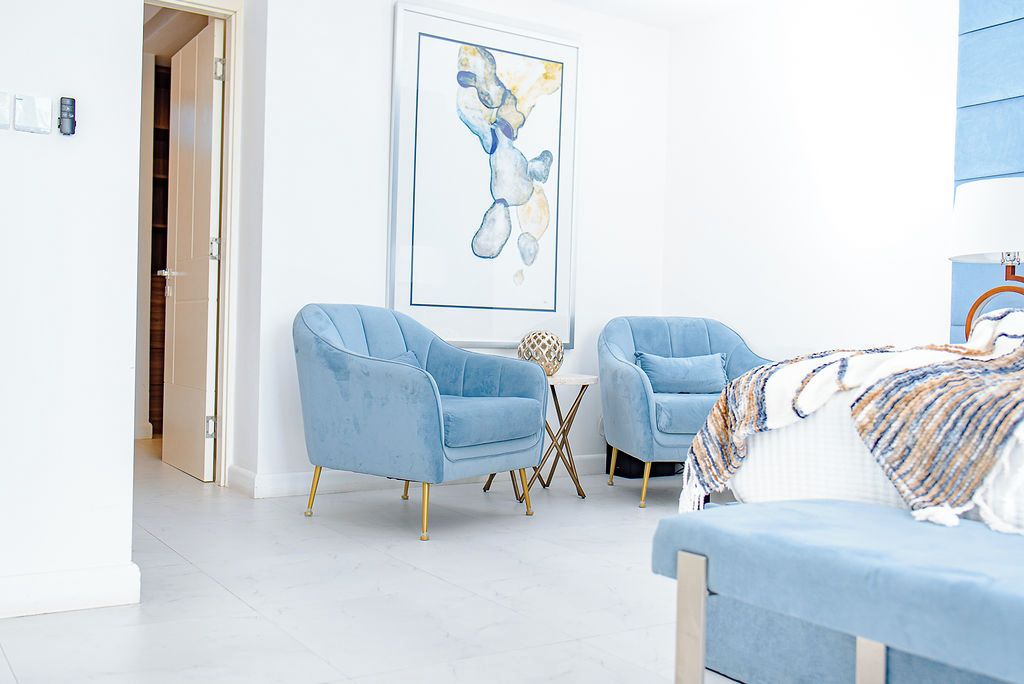 A modern bedroom with two blue armchairs, a bed, and a framed abstract painting on the wall.