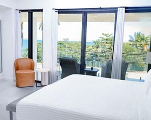 Une chambre d’hôtel avec un grand lit, une chaise, une petite table et un balcon offrant une vue sur l’océan et les palmiers.
