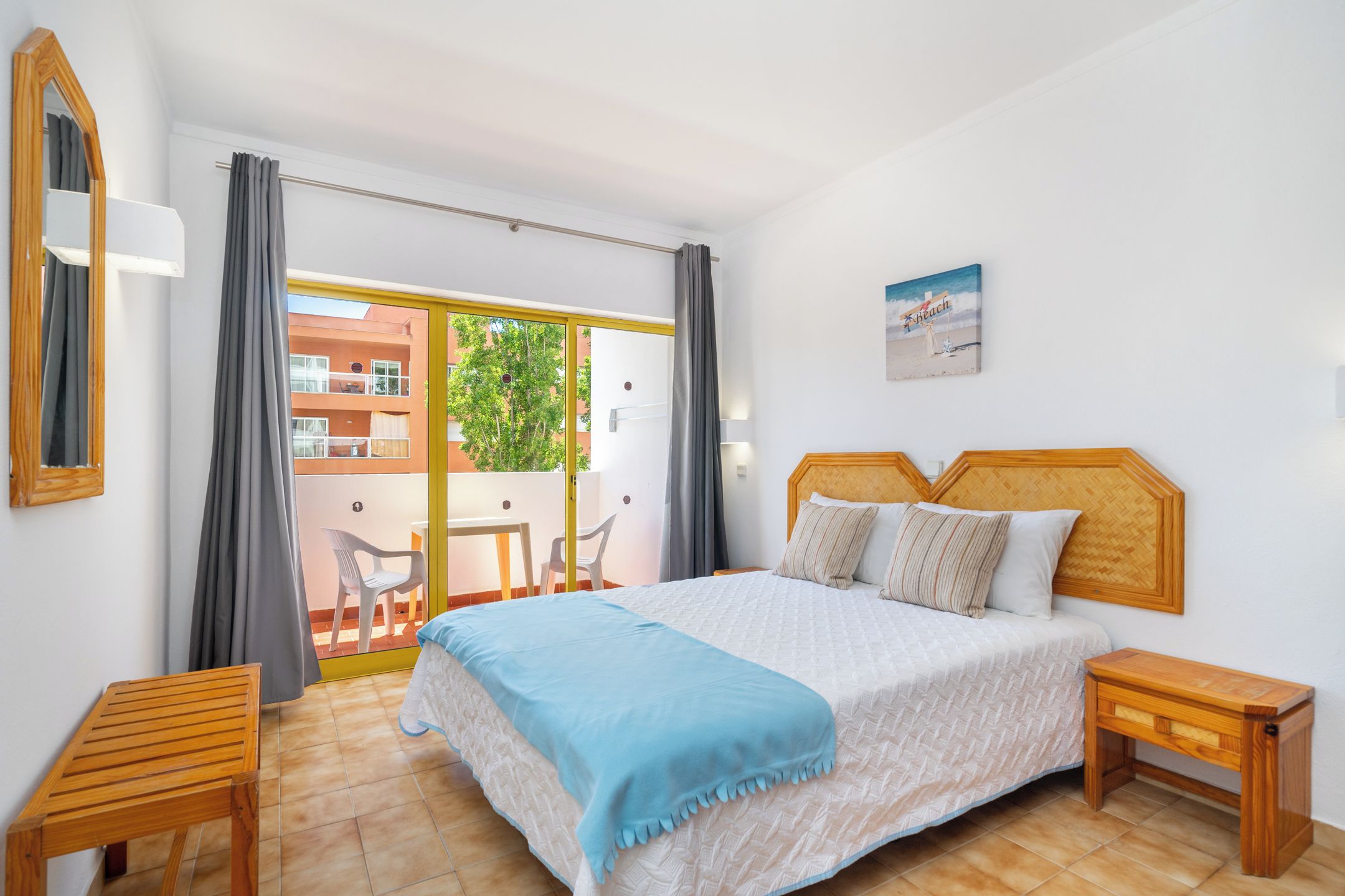 A bedroom with a bed, bedside tables, a balcony with chairs, and a view of buildings outside.