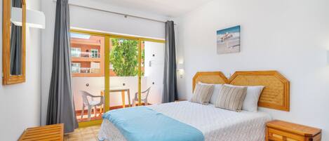 A bedroom with a bed, bedside tables, a balcony with chairs, and a view of buildings outside.