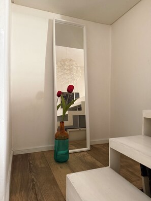 Ein Zimmer mit weißer Wand, Holzboden, einer weißen Bank und einer türkisfarbenen Vase mit roten Blumen.