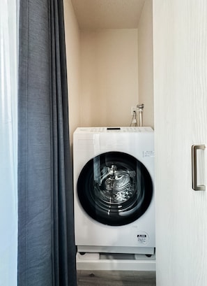 A compact, white front-loading washing machine in a small laundry room with a wooden floor and a door on the right. - HANASTAY MINE (Osaka)