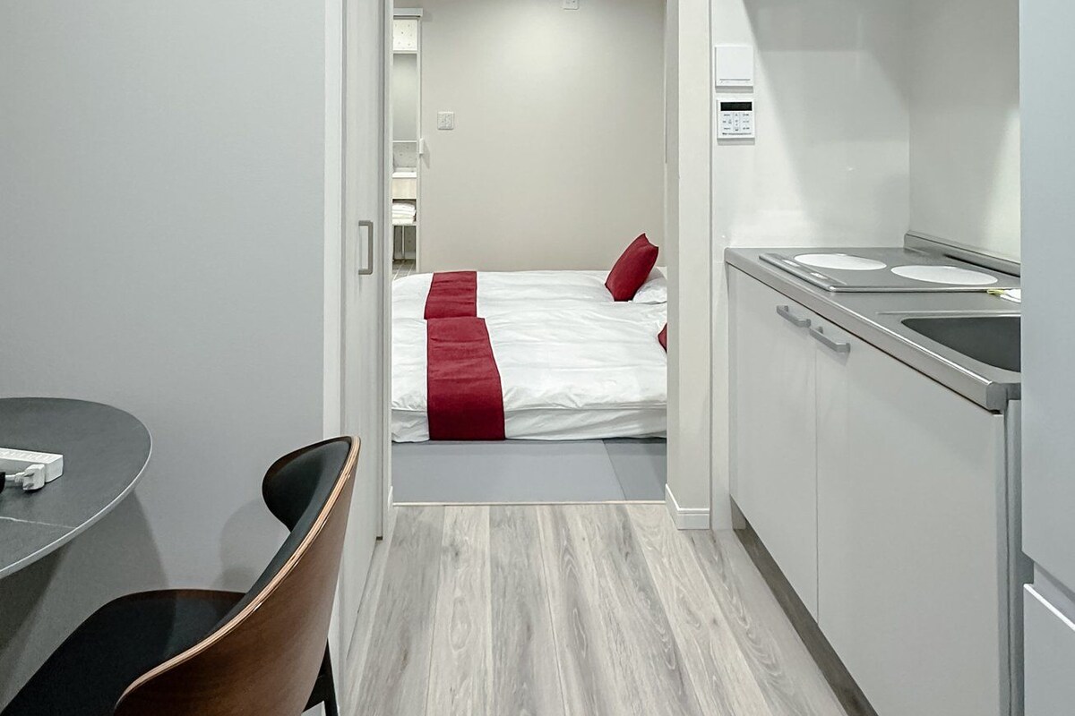 A modern hotel room with a kitchenette, a dining area with a table and chairs, and a bed with red bedding.