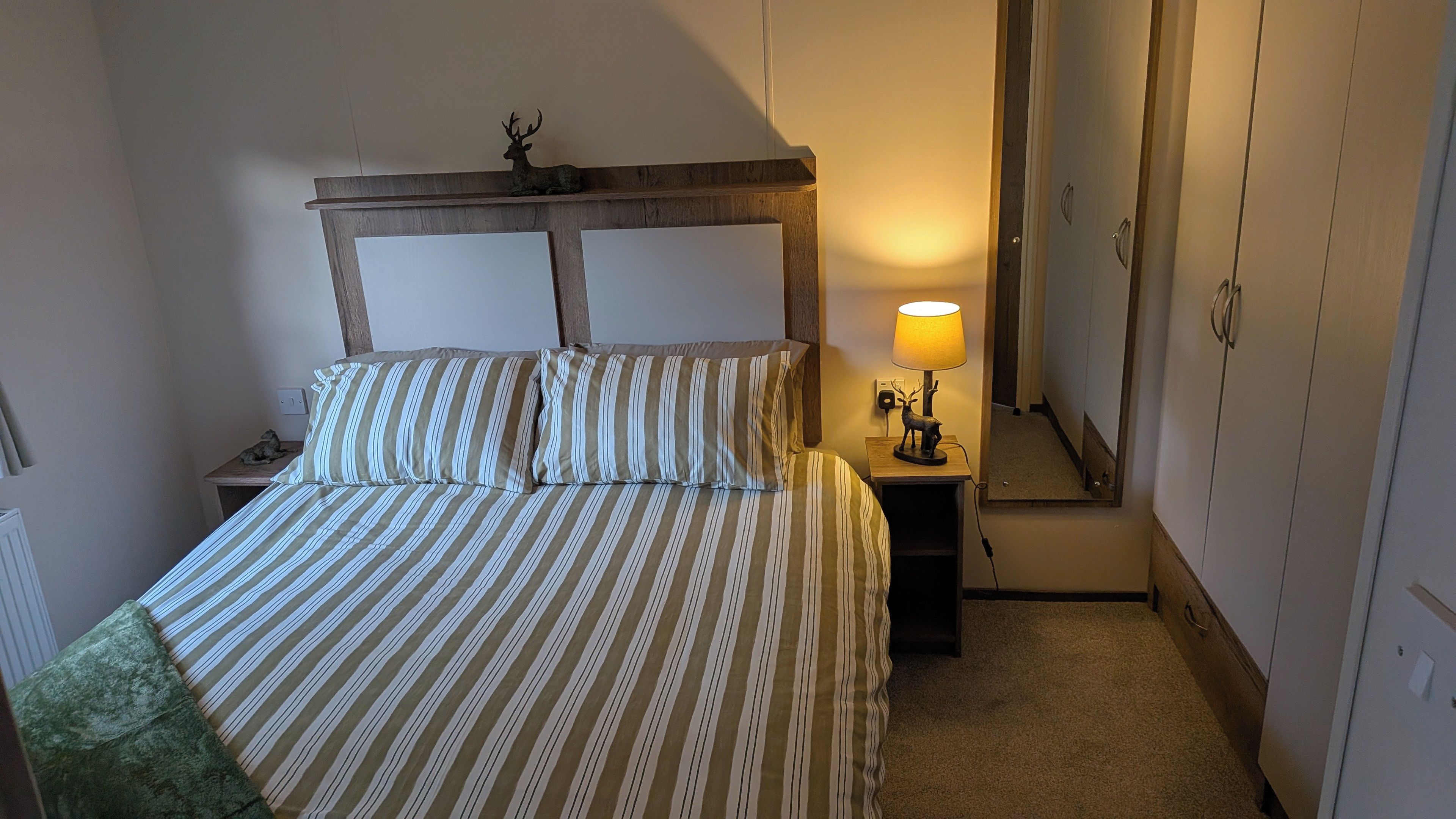 Een bed met gestreepte lakens, een nachtkastje met een lamp en een hertenbeeldje, en een kledingkast in een hotelkamer.