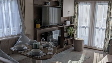 Una sala de estar moderna con un televisor de pantalla plana, un sofá, una mesa de comedor preparada para cuatro personas y un ventanal con cortinas ligeras.