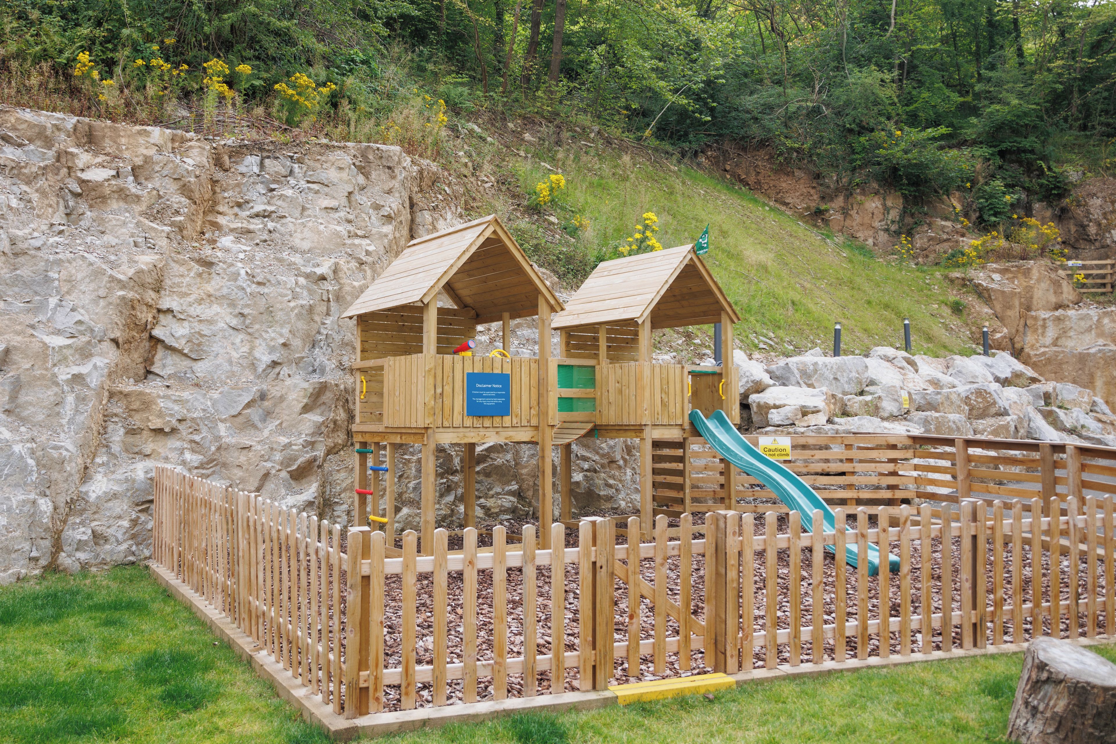 A wooden play structure with a slide, surrounded by a wooden fence, set against a rocky hillside.