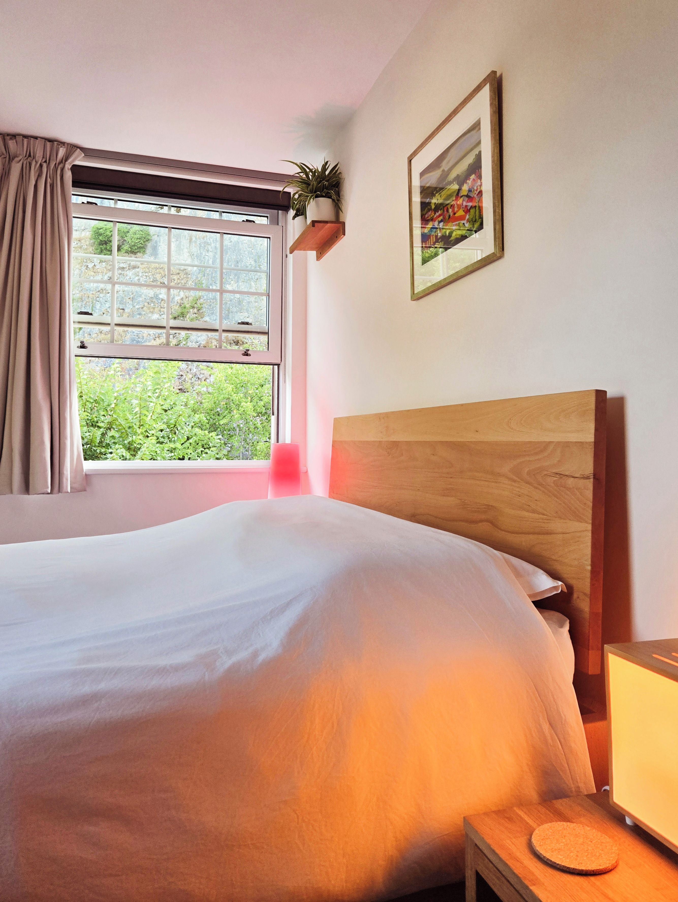 Un dormitorio con cama, ventana con cortinas, mesita de noche con lámpara y un cuadro enmarcado en la pared.
