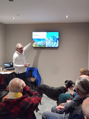 A person presenting in front of a screen displaying a landscape image, with seated individuals and a dog in the room. - Rock Mill Retreat (Stoney Middleton)